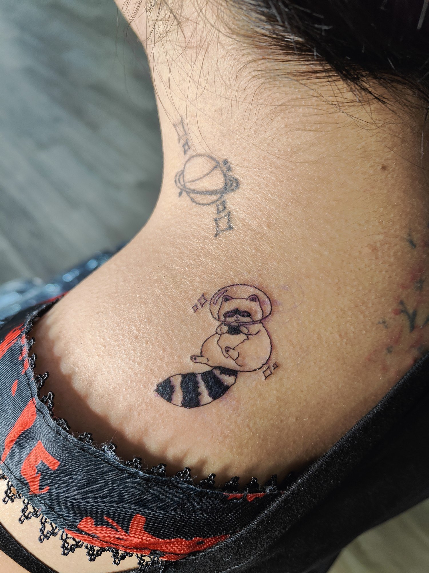 A small seated raccoon wearing a space helmet, with a striped tail and tiny stars on the back of the neck. Tattoo by Edboi