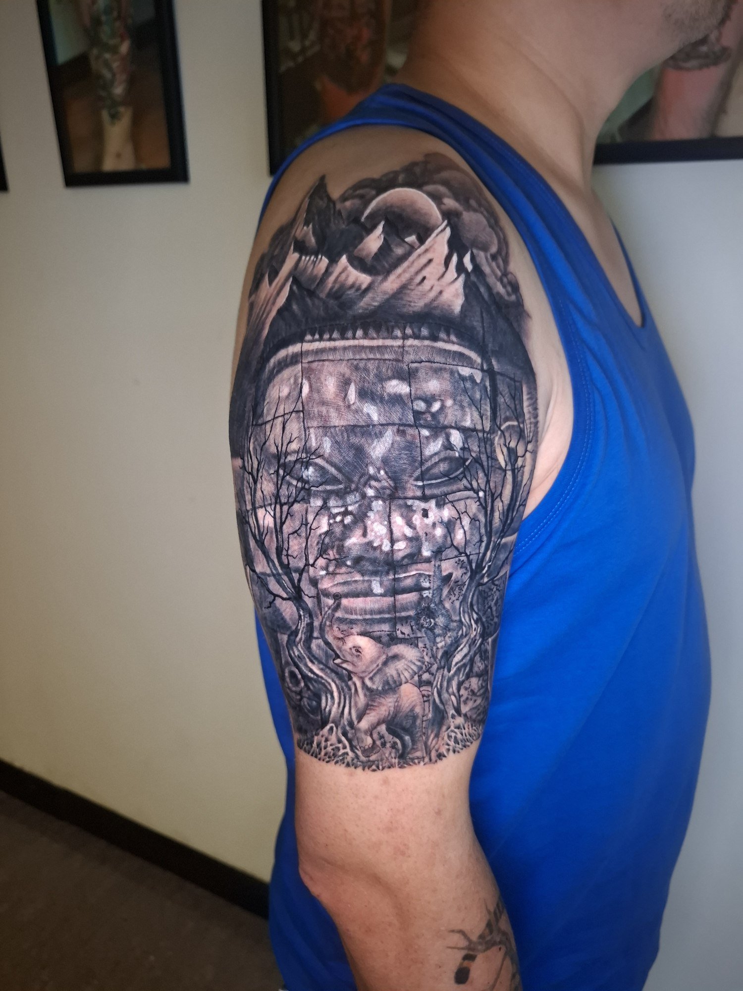 A large stone temple face surrounded by trees, mountains, a moon and an elephant on the shoulder and upper arm. Tattoo by Edboi