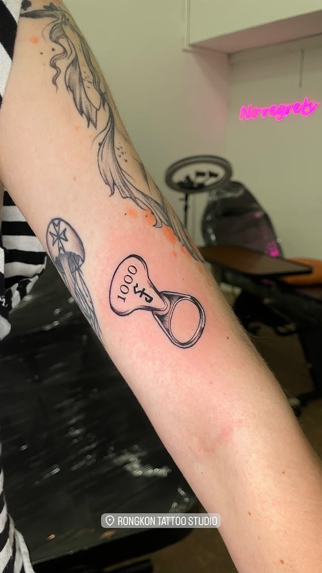 A bottle cap marked 1000 with a small symbol, drawn in black linework on the inner arm. Tattoo by Edboi