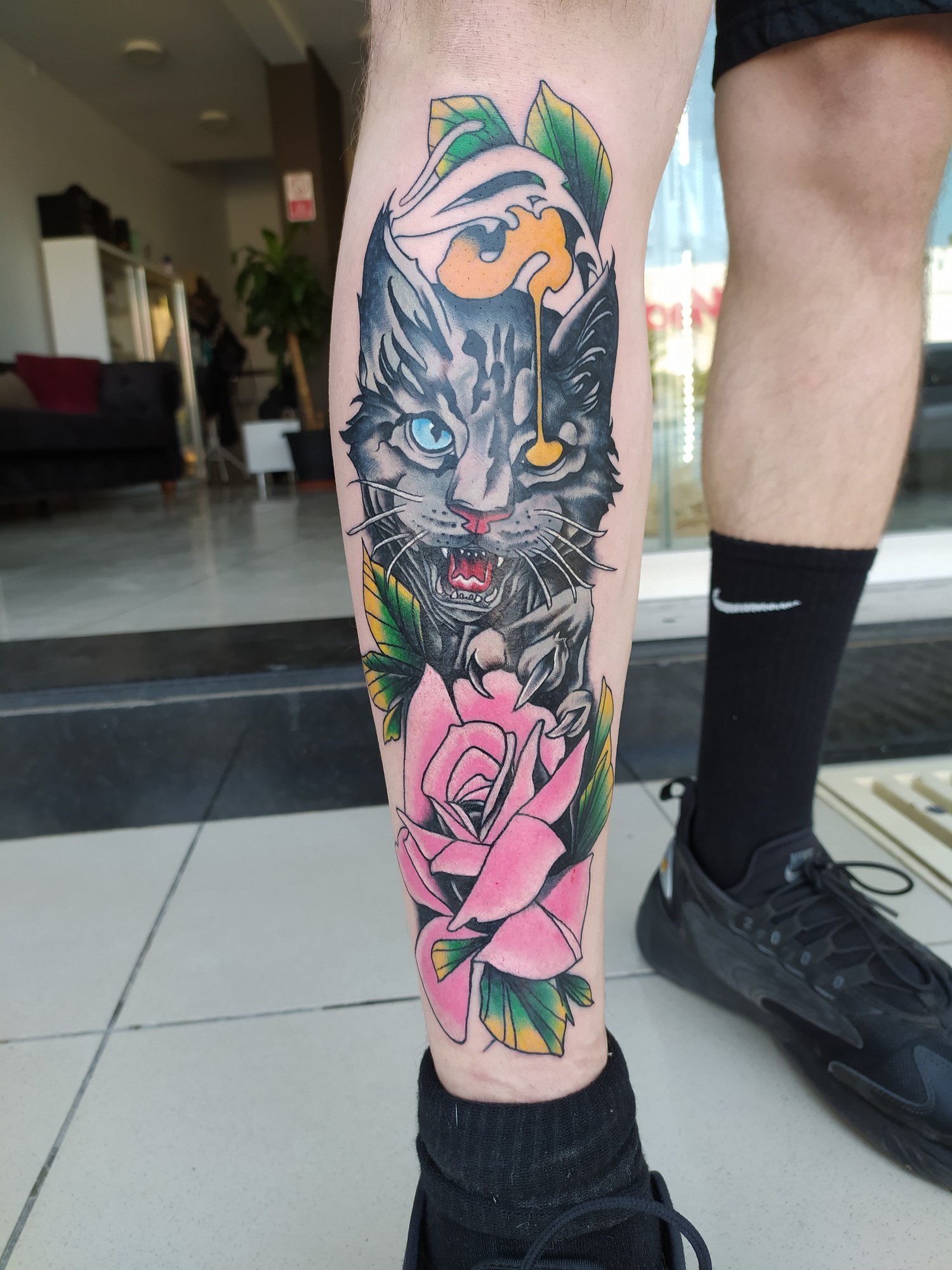 A large stylised cat with a blue eye, a pink rose and green leaves on the lower leg. Tattoo by Edboi