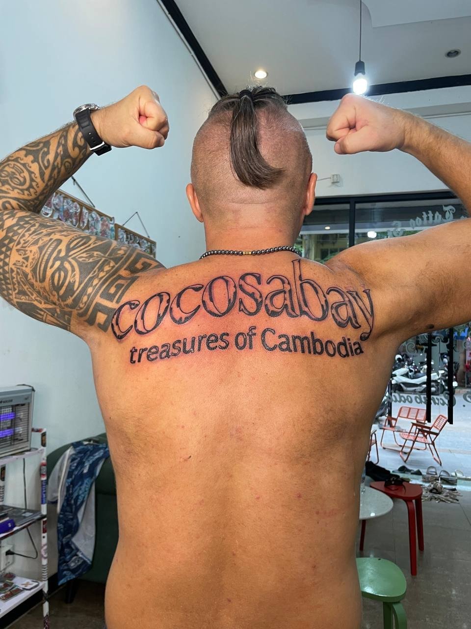 Large outlined Cocosabay lettering with a second line reading treasures of Cambodia across the upper back. Tattoo by Edboi