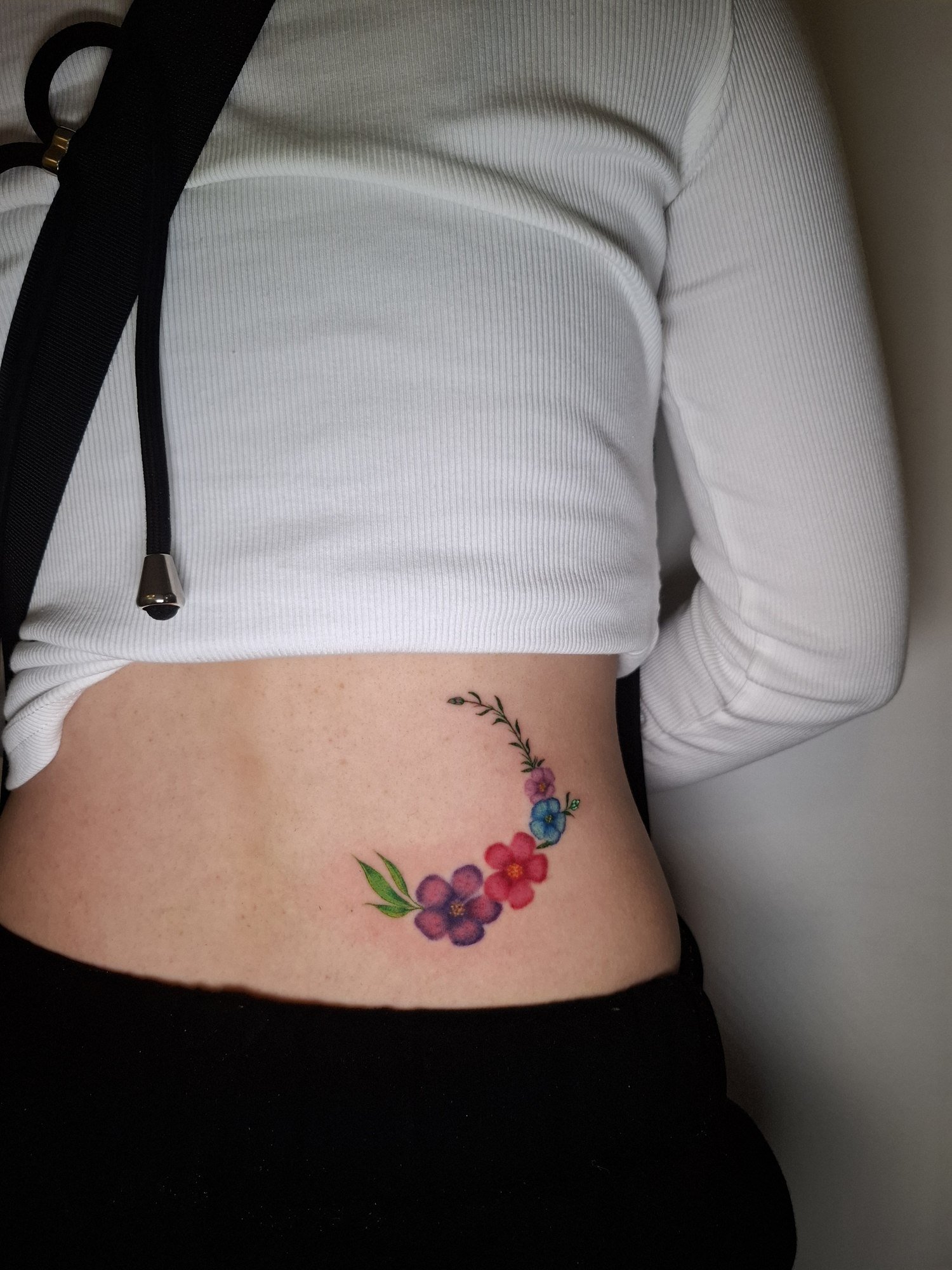 Purple, pink and blue flowers with green leaves arranged in a crescent on the side of the waist. Tattoo by Edboi