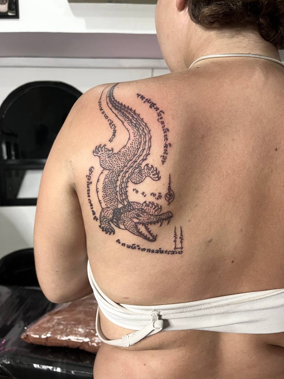 A detailed crocodile surrounded by ornamental lettering and fine symbols on the upper back. Tattoo by Edboi