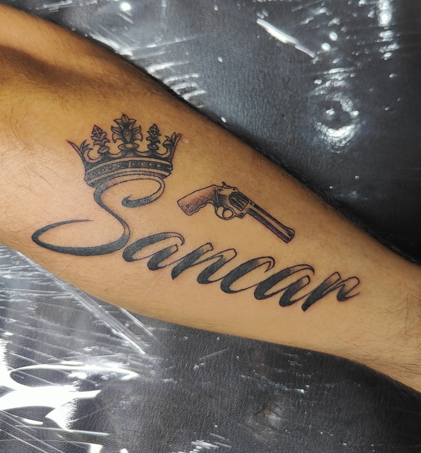 Large flowing name lettering with a crown and a small revolver motif. Tattoo by Edboi