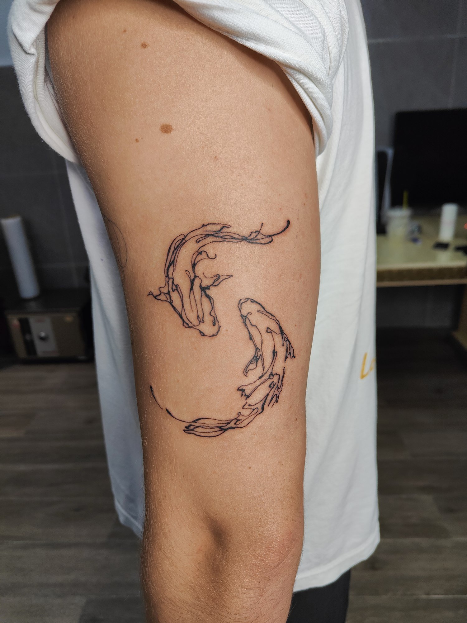 Two fish arranged in a circle with flowing fins and delicate abstract black linework on the upper arm. Tattoo by Edboi