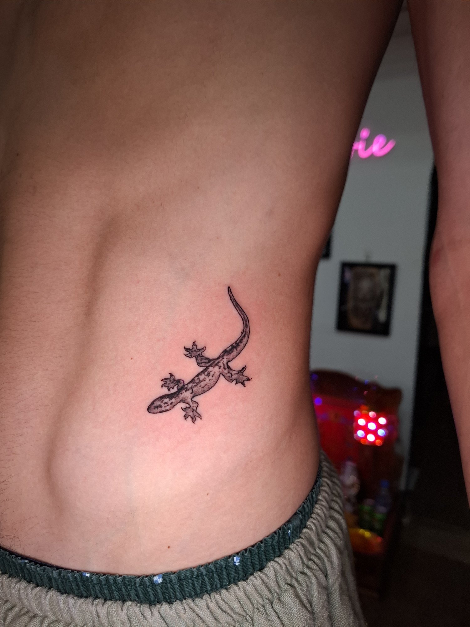 A small gecko with mottled shading and a curved tail on the side of the torso. Tattoo by Edboi