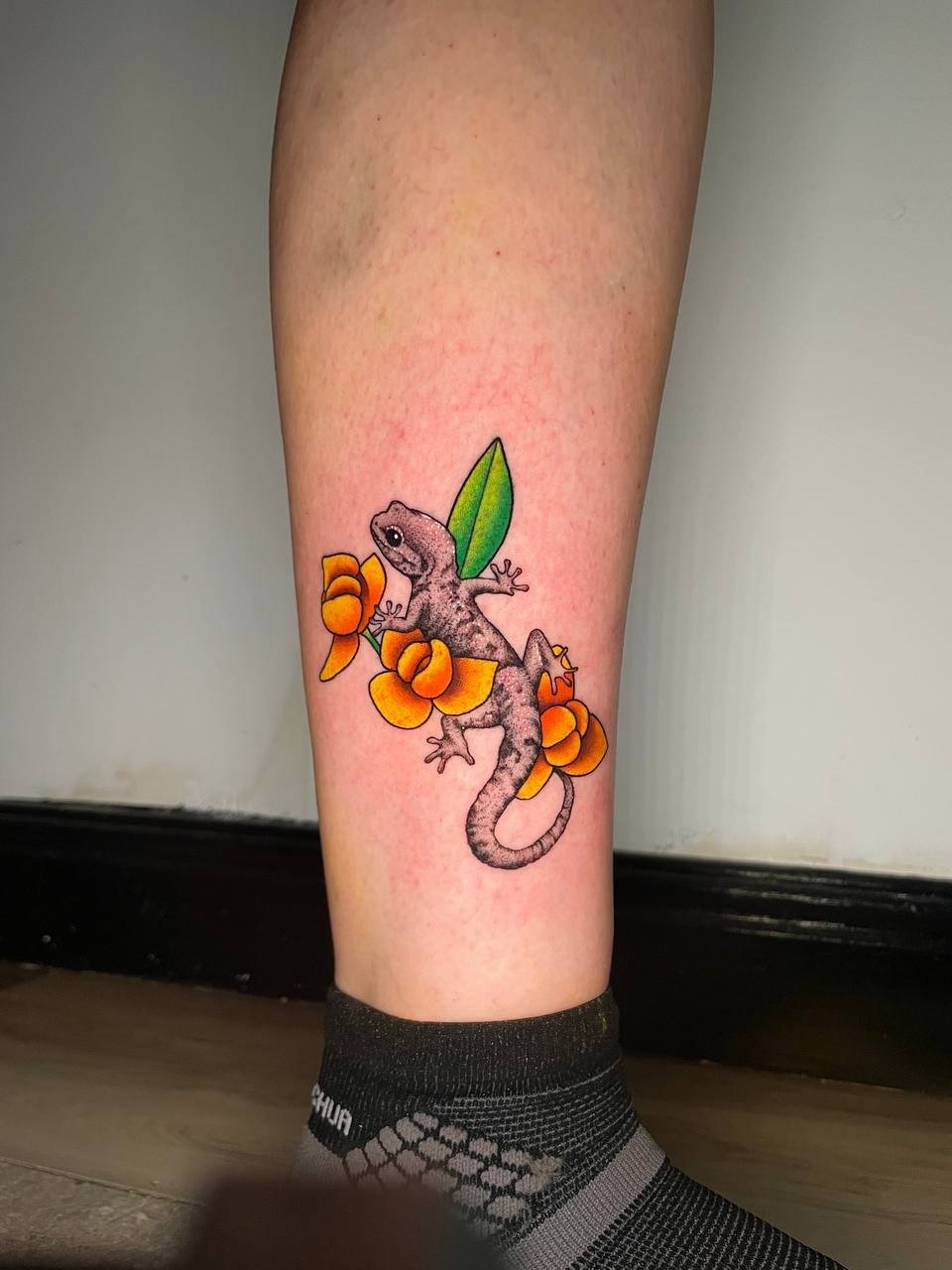 A shaded gecko among yellow flowers and a green leaf on the lower leg. Tattoo by Edboi