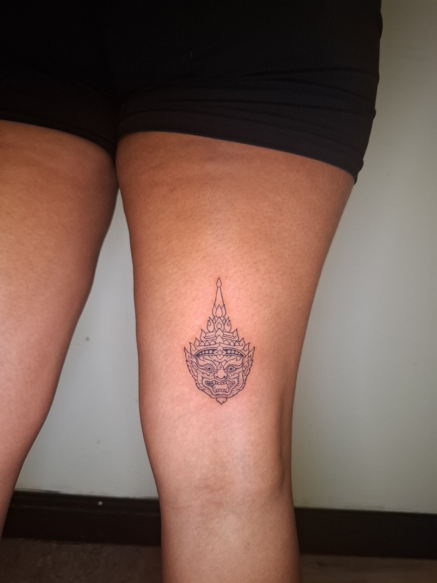 A small symmetrical ornamental face with an elaborate pointed headdress on the thigh. Tattoo by Edboi