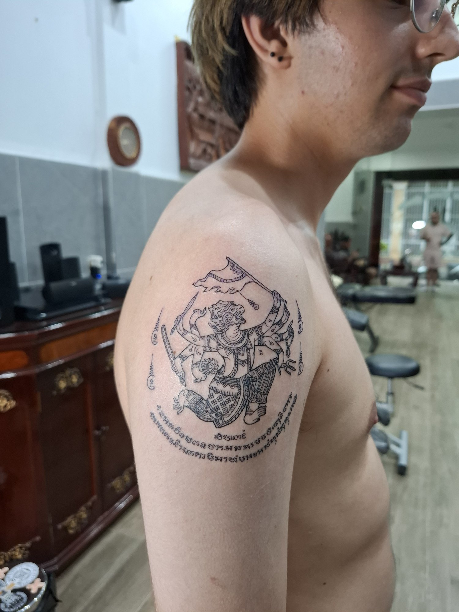 An intricately outlined guardian figure with a flag, ornamental details and surrounding script on the shoulder. Tattoo by Edboi