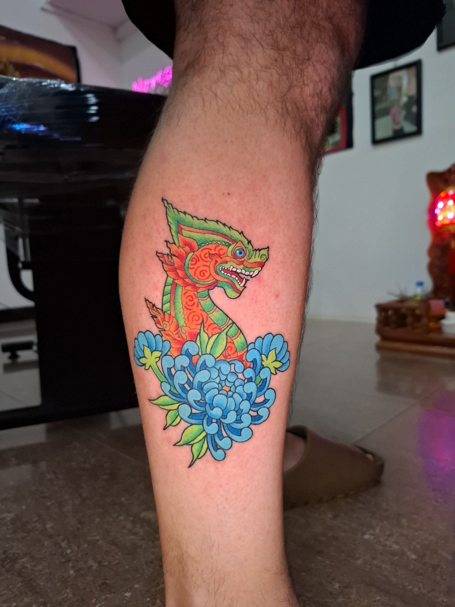 A colourful ornamental Naga with green and orange details rising above blue flowers on the calf. Tattoo by Edboi