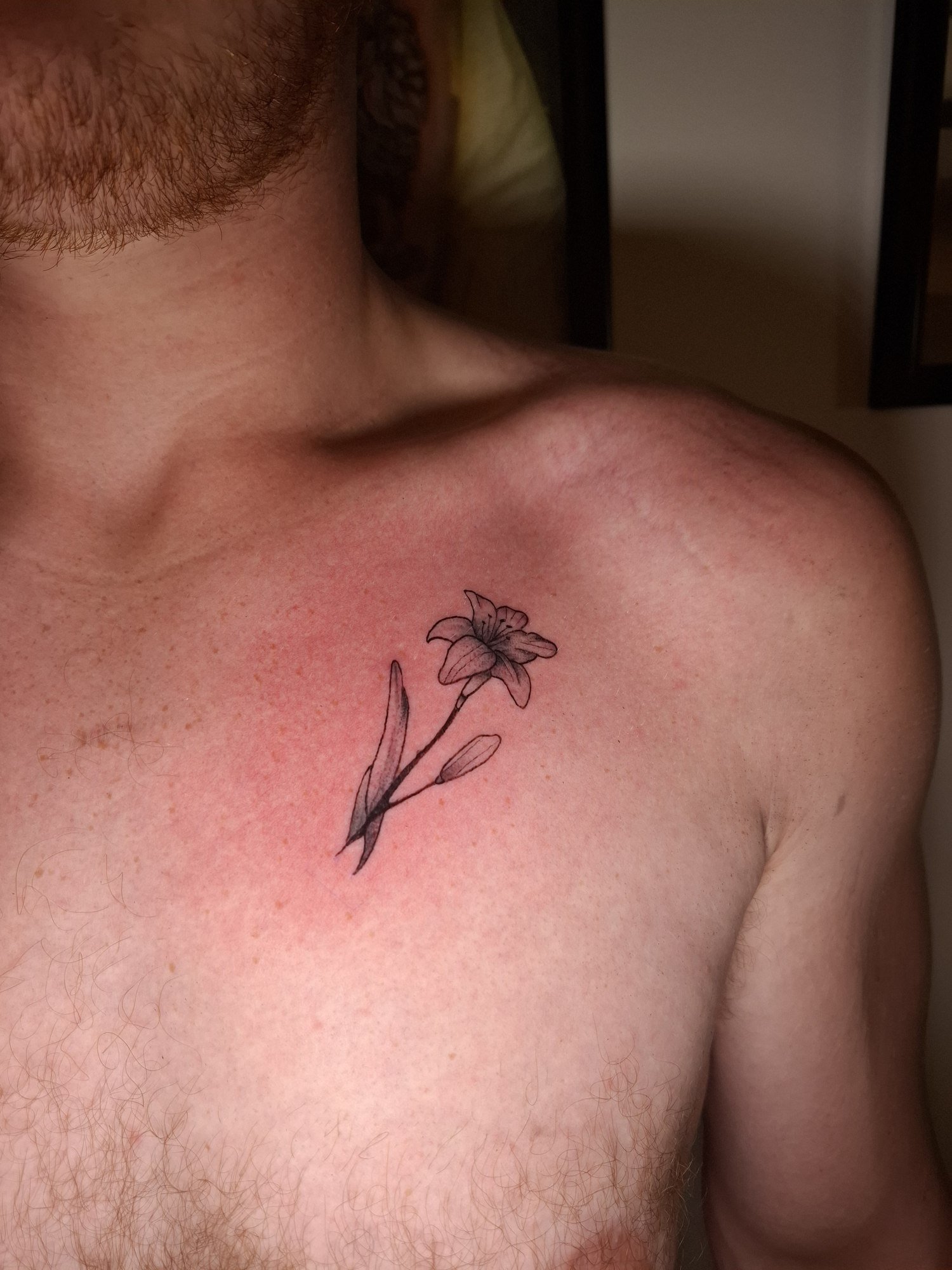 A small lily with a bud, slender stem and softly shaded leaves on the upper chest. Tattoo by Edboi