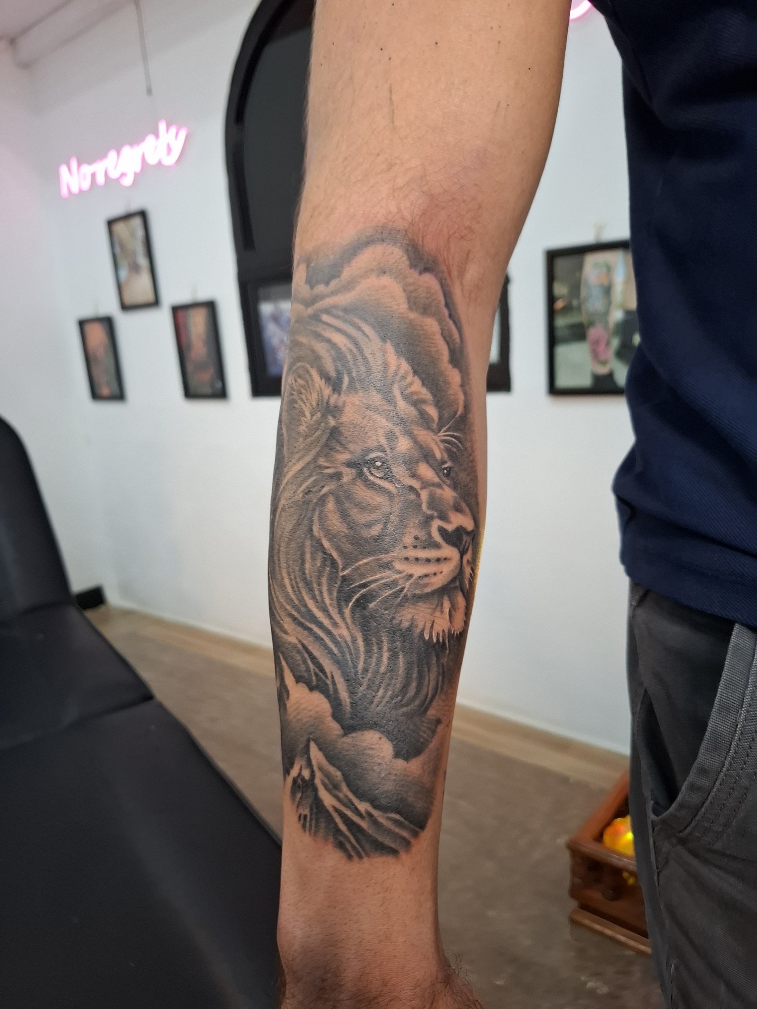 A shaded lion portrait framed by clouds and mountain peaks on the forearm. Tattoo by Edboi