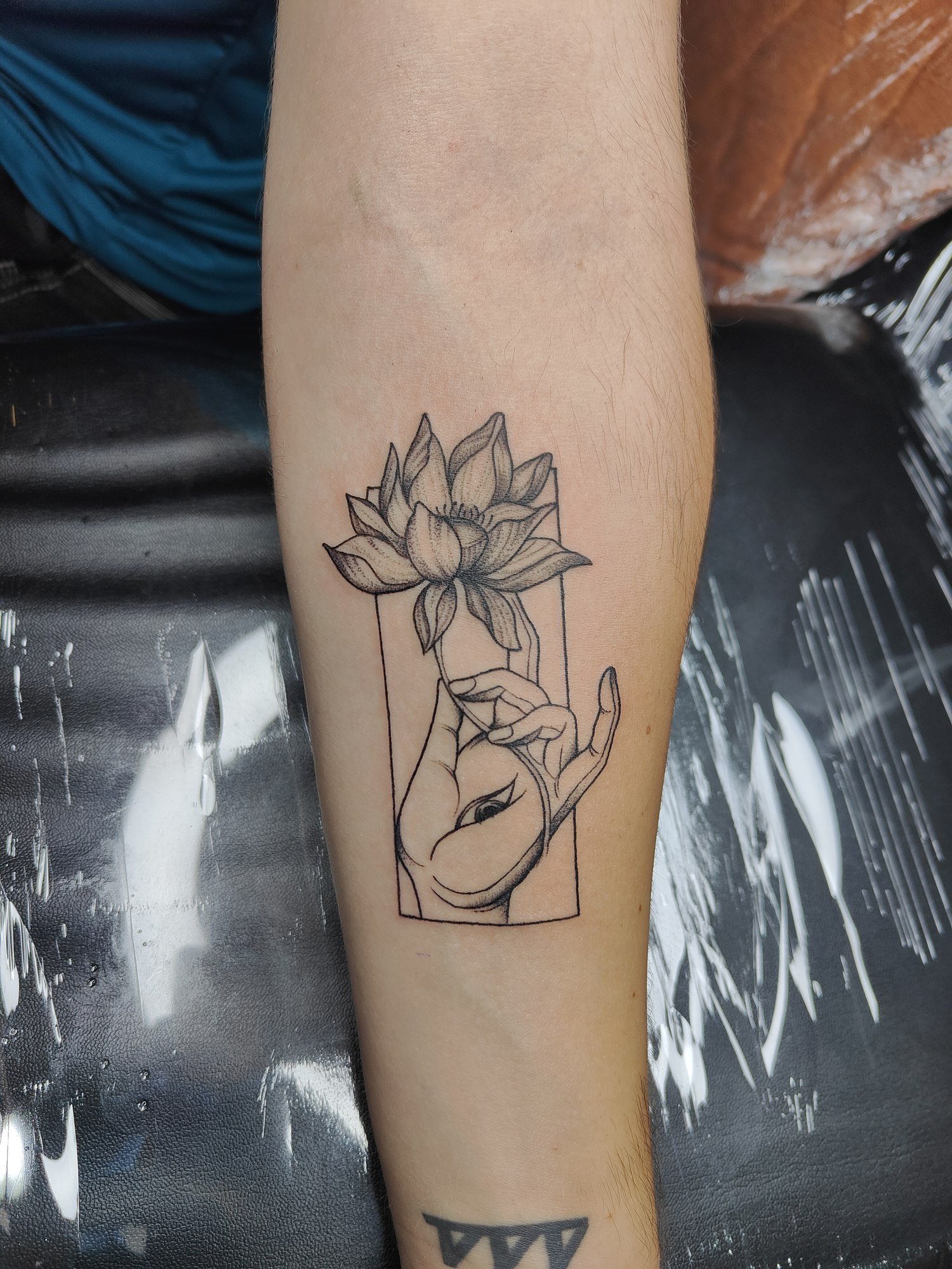 A linework hand holding a shaded lotus within a rectangular frame on the forearm. Tattoo by Edboi