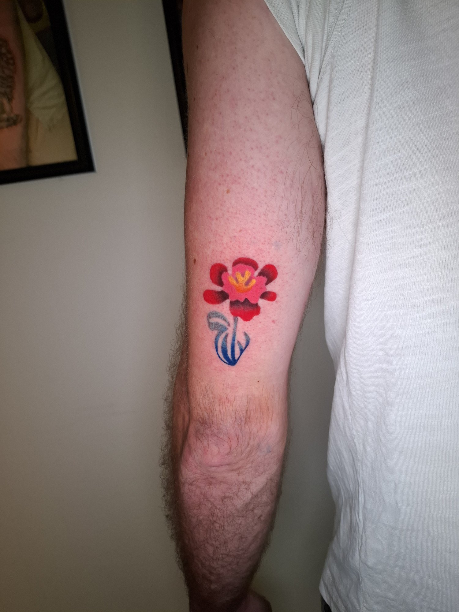 A stylised red flower with a yellow centre and flowing blue stem on the arm. Tattoo by Edboi