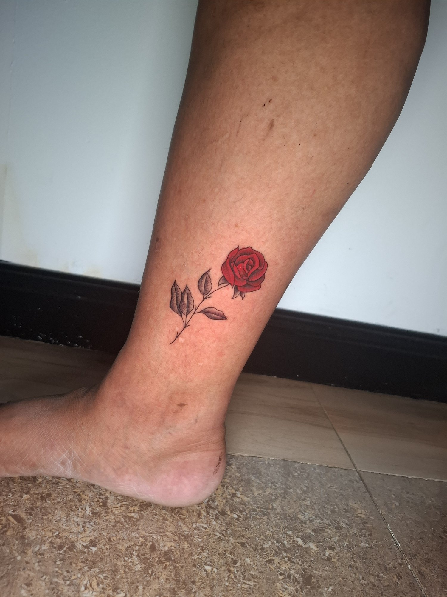 A small red rose with a fine stem and shaded black and grey leaves above the ankle. Tattoo by Edboi