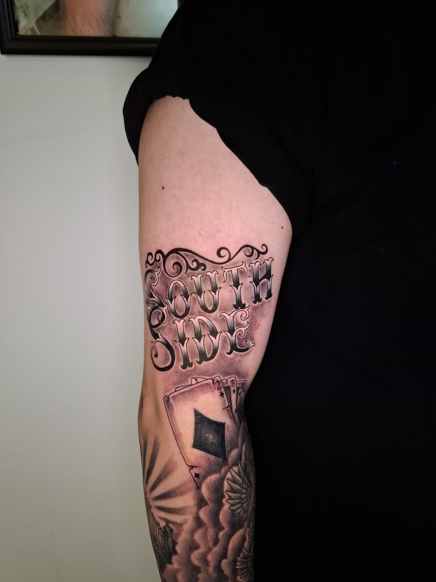 Large shaded gothic lettering with ornamental flourishes above playing cards on the upper arm. Tattoo by Edboi